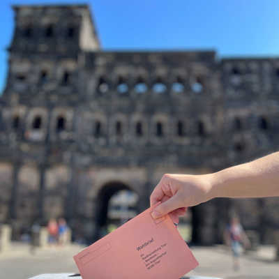 Ein Umschlag wird in eine Urne gesteckt, im Hintergrund sieht man verschwommen die Porta Nigra