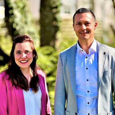 Die Amtsleiterin der Wirtschaftsförderung Trier Nina Womelsdorf und der stellvertretende Amtsleiter Eric Krischel stehen nebeneinander vor grünen Bäumen und lächeln in die Kamera.