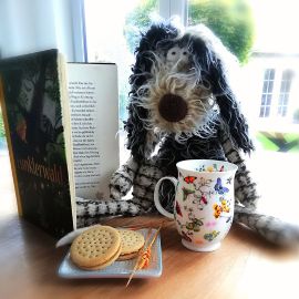 Ein zotteliger Hund aus Stoff mit einer Tasse, Keksen und einem aufgeschlagenen Buch