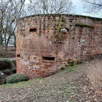 Eine rundförmige, aus roten Sandsteinblöcjken gemauerte historische Wehranlage mit Schießscharten steht in einem Park.