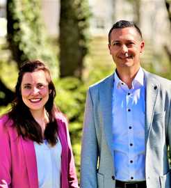 Die Amtsleiterin der Wirtschaftsförderung Trier Nina Womelsdorf und der stellvertretende Amtsleiter Eric Krischel stehen nebeneinander vor grünen Bäumen und lächeln in die Kamera.
