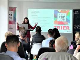Die Amtsleiterin der Wirtschaft hält einen Vortrag vor Mitgliedern des Netzwerks "Attraktive Unternehmen Trier". Sie steht vor einer Projektion des Netzwerklogos.