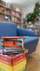 Ein Stapel Bücher (Das Tagebuch der Anne Frank, The Hunger Games, Harry Potter, Call me by your Name, Das Schicksal ist ein mieser Verräter, Der Report der Magd) auf einem Hocker vor verschwommenem Hintergrund (Regale der Bücherei und blaues Sofa)