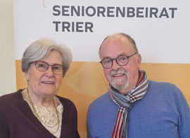 Beide Personen lächeln in die Kamera vor einem Aufsteller mit der Aufschrift "Seniorenbeirat Trier"